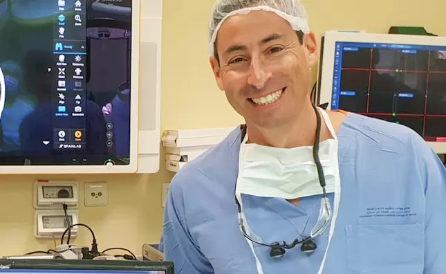 Dr. Amir Kershenovich wearing surgical scrubs and a mask around his neck, smiling next to medical monitors in the OR